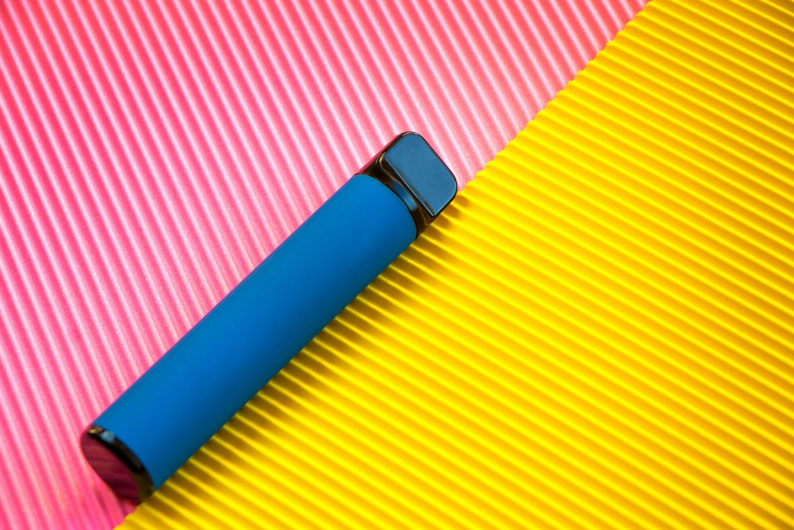 Ein blaues Einweg-E-Zigaretten-Gerät liegt auf einem bunten Hintergrund, der aus diagonal gestreiften pinken und gelben Abschnitten besteht.