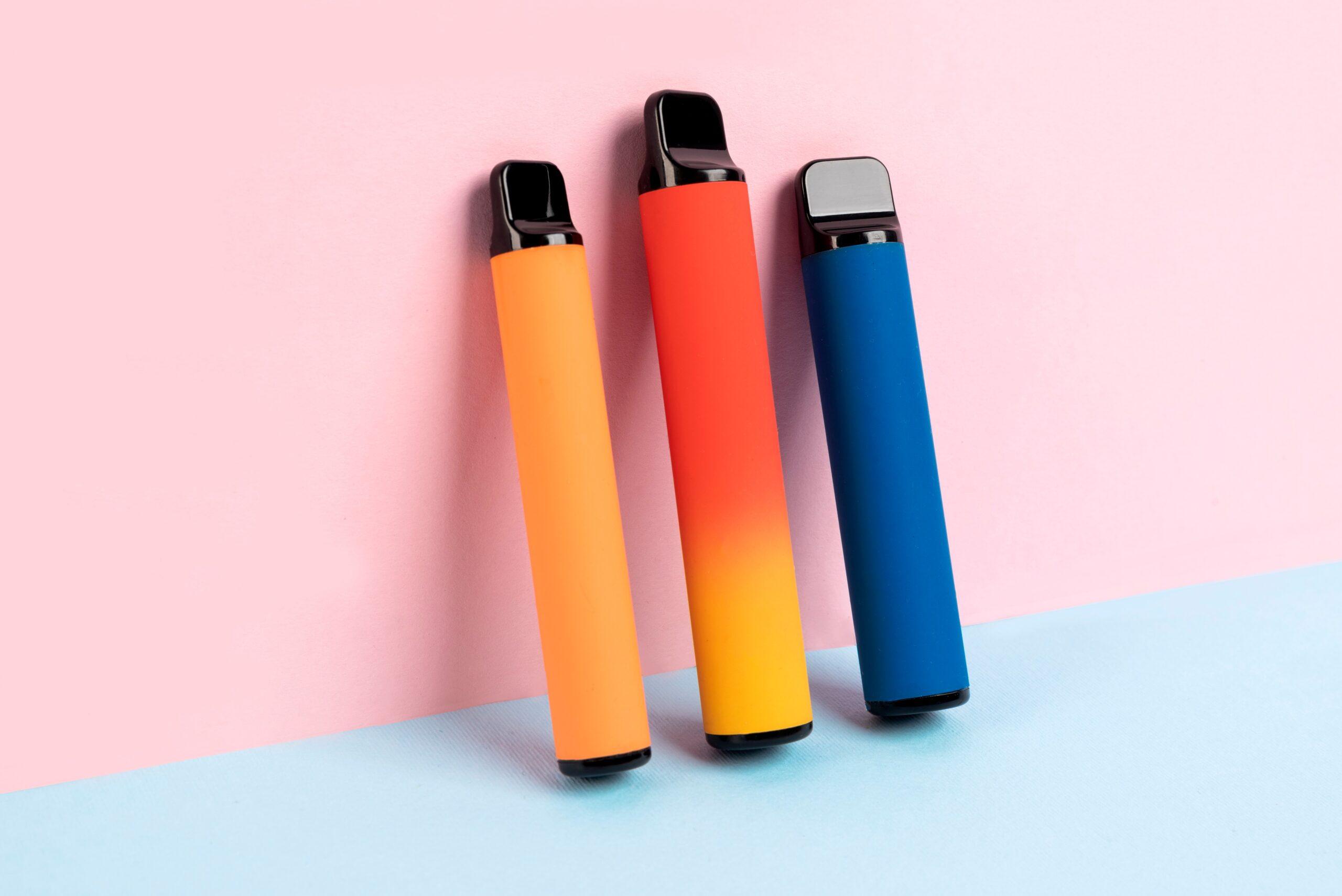 Drei Einweg-E-Zigaretten in den Farben Orange, Rot-Gelb und Blau stehen aufrecht auf einem zweifarbigen Hintergrund in Pastellrosa und Pastellblau.
